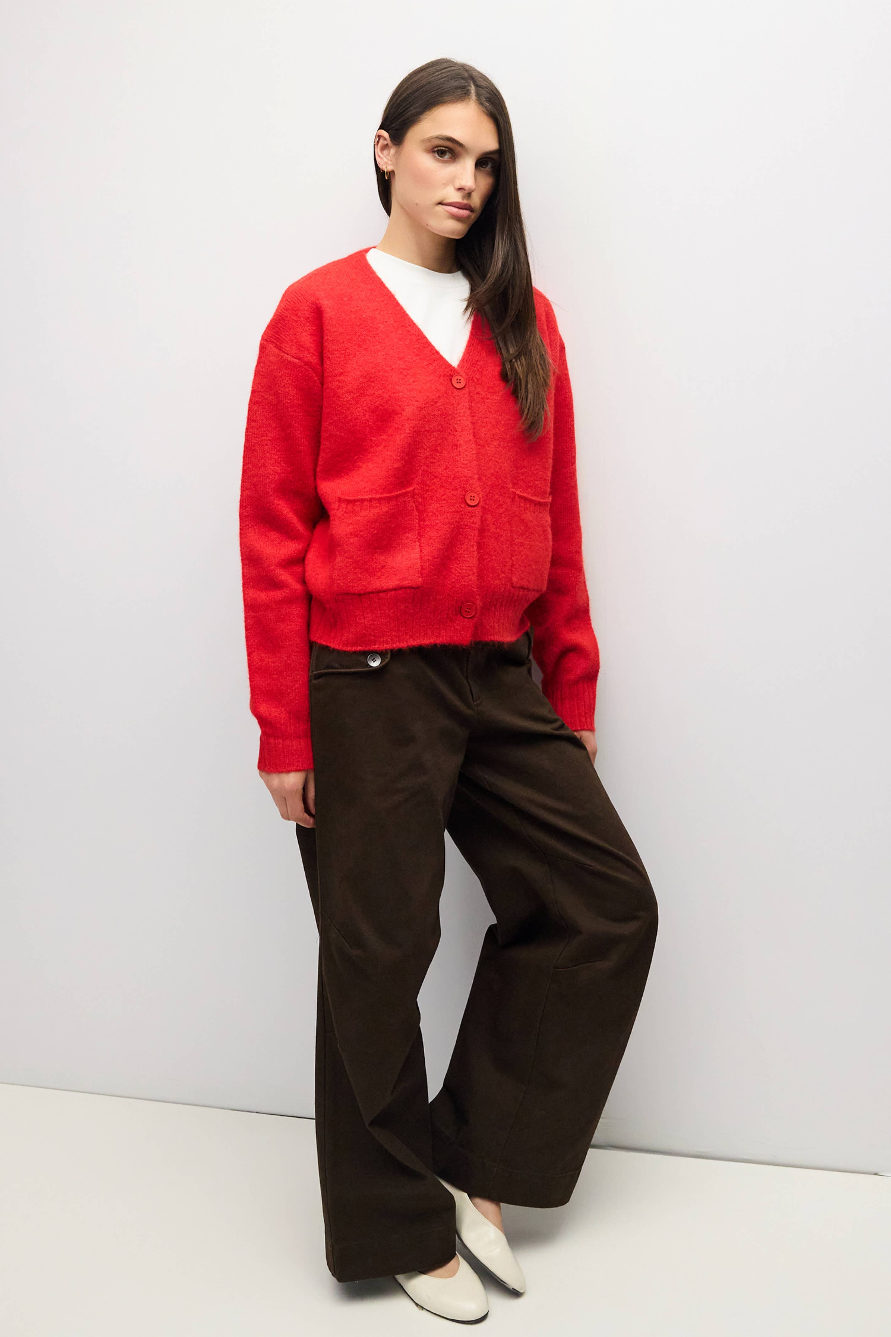 A woman modeling a bright red knit cardigan sweater with front buttons and patch pockets, paired with dark brown wide-leg trousers and white pointed flats, standing against a plain light gray studio wall.