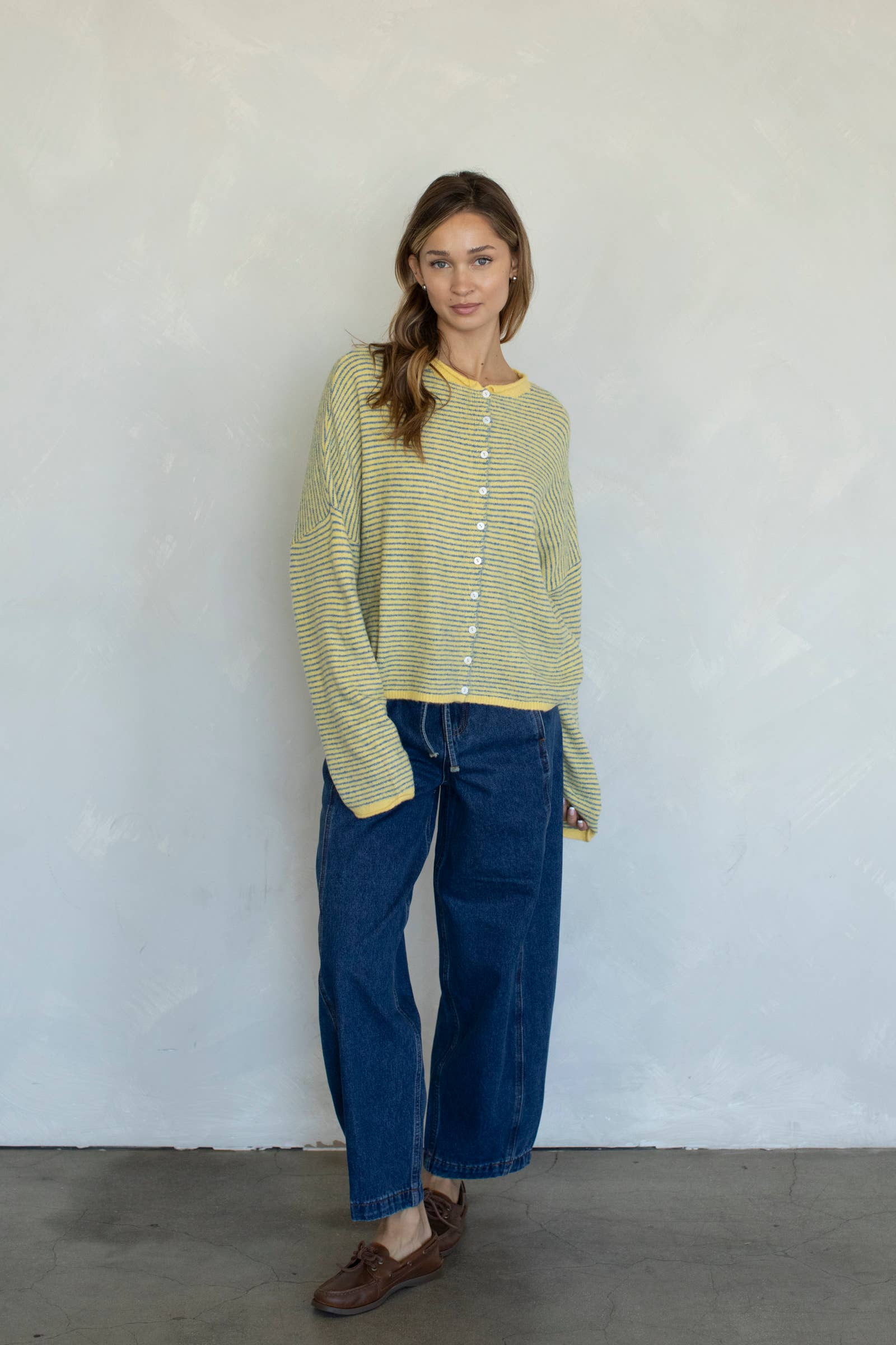 Woman wearing a green striped shirt and blue jeans standing against a white wall.