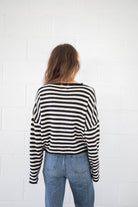 Woman wearing blue jeans and button down, black and white striped, cardigan standing facing away from the camera against white brick black ground 