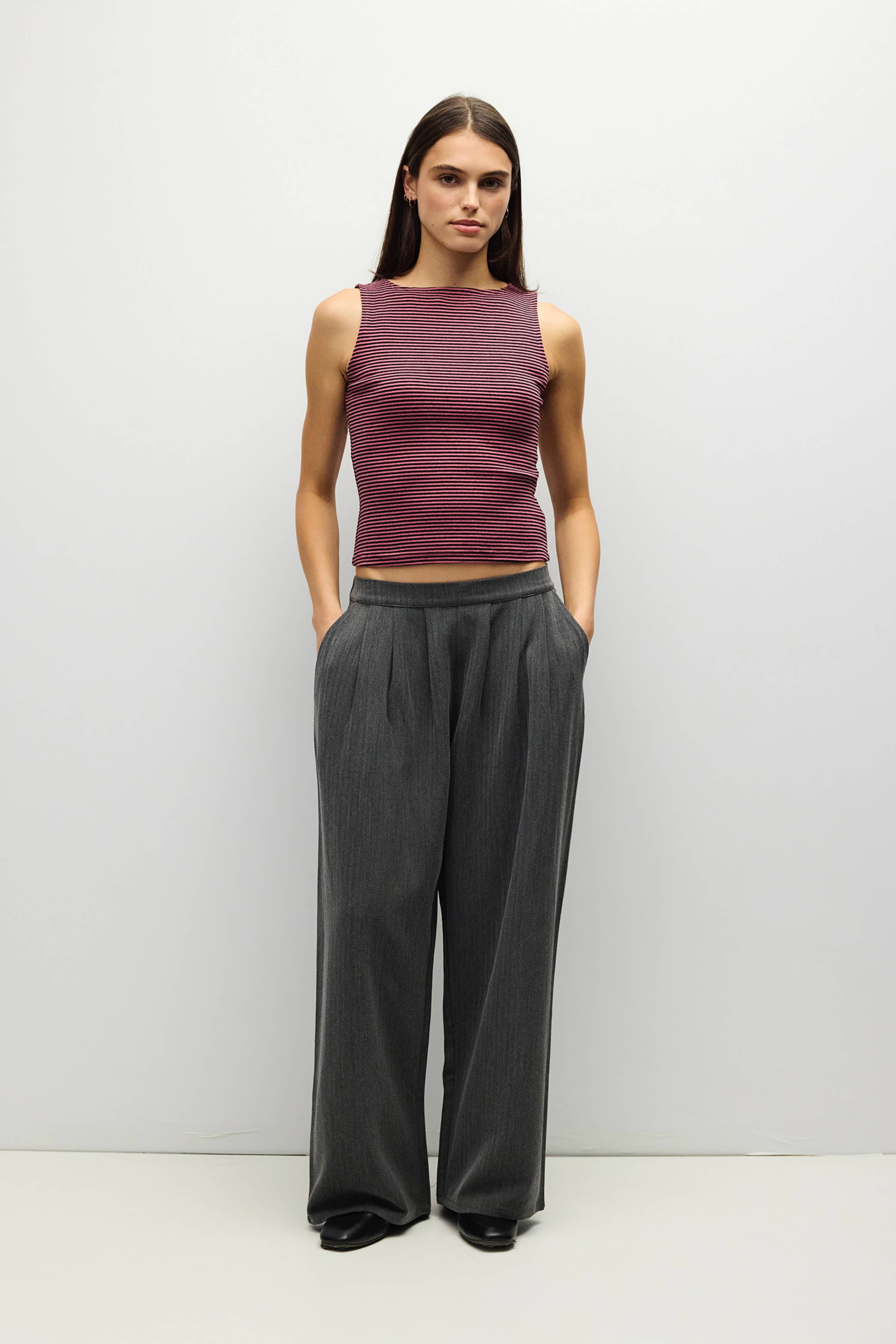 A woman stands against a plain light gray studio wall wearing a sleeveless heathered plum crop top and dark charcoal wide-leg trousers.