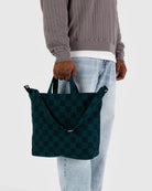 Model standing against white background holding BAGGU square navy and green checkered tote bag with handles and adjustable straps 