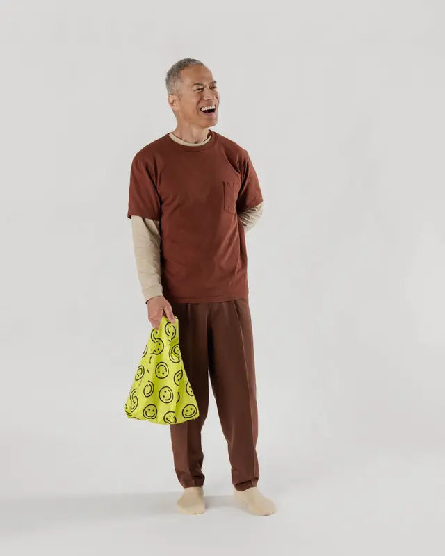 Model against white background wearing orange and brown outfit hold neon yellow smiley face reusable BAGGU bag