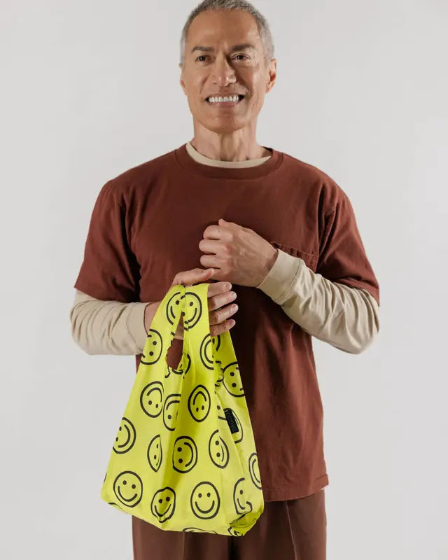 Model against white background wearing orange and brown outfit hold neon yellow smiley face reusable BAGGU bag