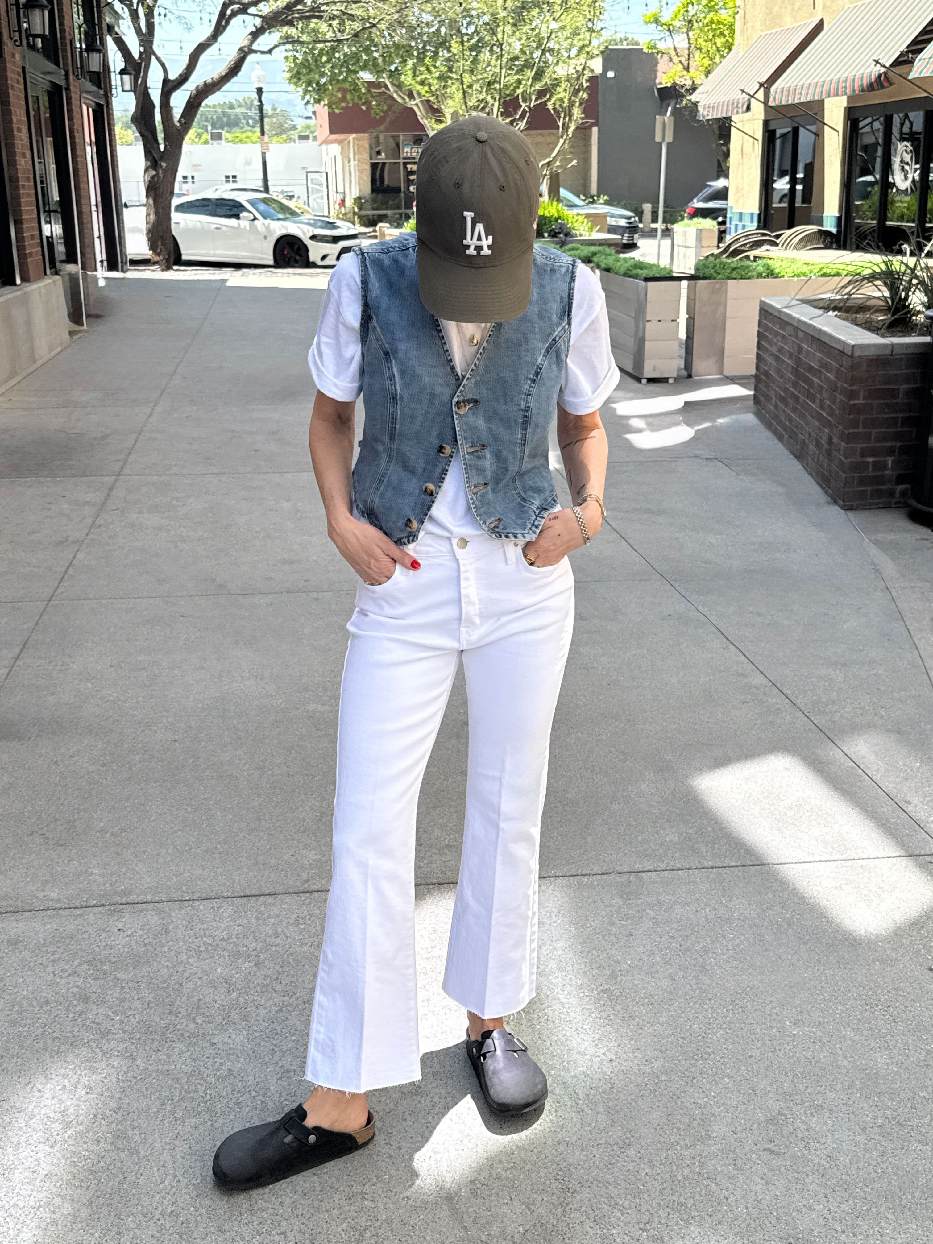 Person wearing a denim vest, white shirt, white pants, and black shoes on a sidewalk.
